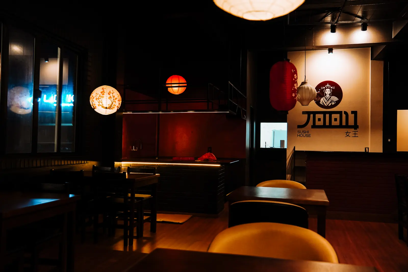 Salão da JOOU à noite, com lanternas japonesas acesas, mesas de madeira e a gueixa do logo na parede